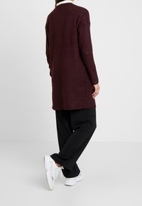 Cárdigan largo burdeos de tejido de punto, con un corte relajado, cuello redondo y puños de canalé, combinado con pantalones negros holgados y zapatillas blancas.