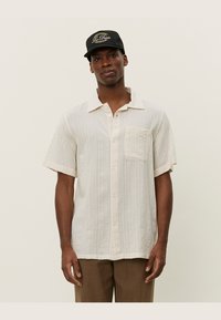 Chemise à manches courtes, de couleur beige clair, avec des rayures texturées et une poche poitrine, assortie d'une casquette noire.