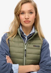 Olivgrön pufferväst med dragkedja och svart logotyp, bärs över en marinblå sweatshirt och en ljusblå randig skjorta.