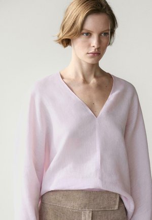 Ung kvinde iført en lyserød V-hals bluse med lange ærmer, stoppet ned i højtaljede beige bukser mod en ensfarvet baggrund.