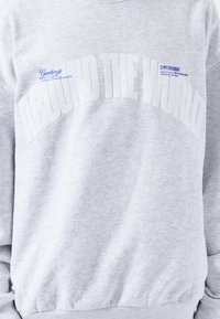Sudadera gris con cuello redondo, que presenta un gran texto blanco "ALREDEDOR DEL MUNDO" y detalles de etiqueta azul. Tejido suave y texturizado.