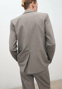 Traje ajustado gris claro con un blazer estructurado y pantalones a juego. La tela tiene una textura sutil y presenta una abertura central en la parte posterior.