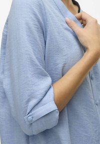 Svetlo modra, lanena srajca s plaščem v obliki črke V, zavihanimi rokavi in subtilno teksturo. Ima okrogle gumbe po sprednji strani.