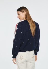 Marineblå tröja med en texturerad yta, dekorerad med silverpaljetter; har sportiga ränder i röd och beige på ärmarna.