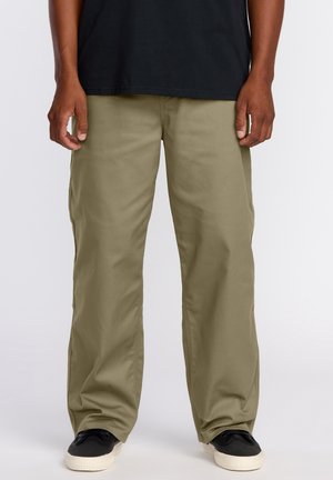 Homme portant un pantalon olive ample, des baskets noires avec des semelles blanches, et un t-shirt noir, debout devant un fond blanc.