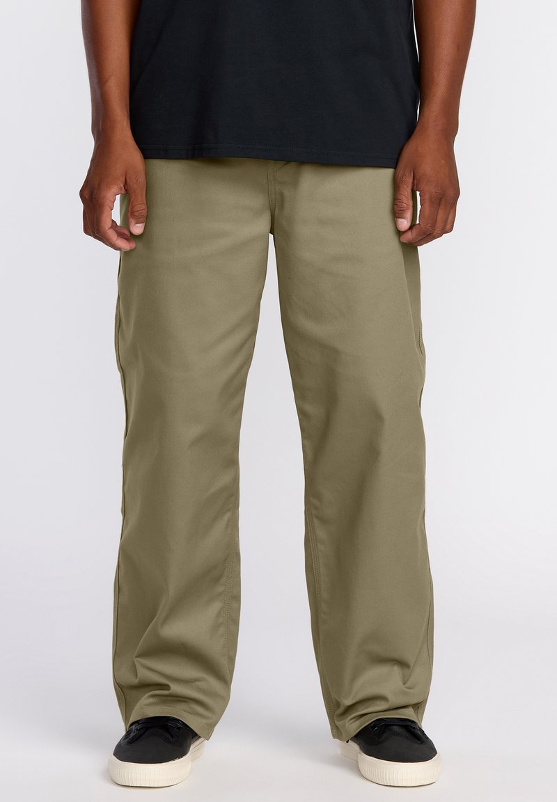 Homme portant un pantalon olive ample, des baskets noires avec des semelles blanches, et un t-shirt noir, debout devant un fond blanc.