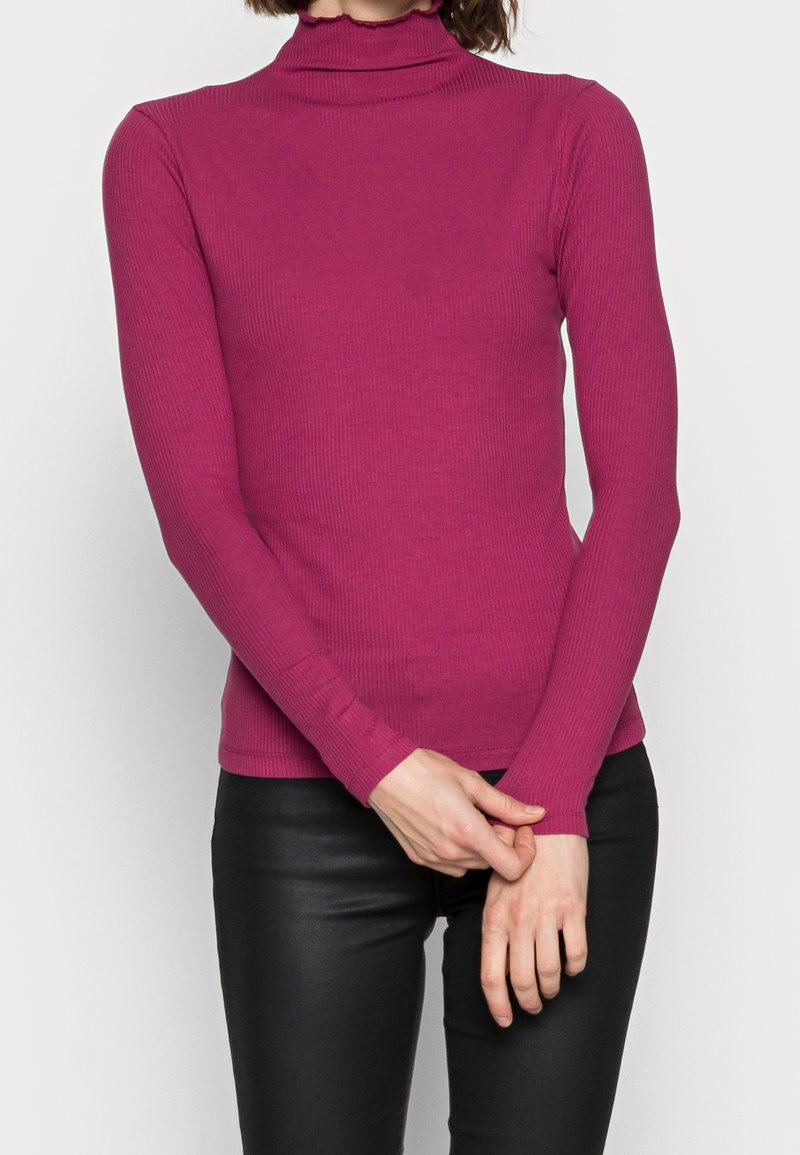 Dopasowana bluzka z golfem w kolorze fuksji, o prążkowanej fakturze, z długimi rękawami i falbanką przy kołnierzu, zestawiona z czarnymi spodniami.