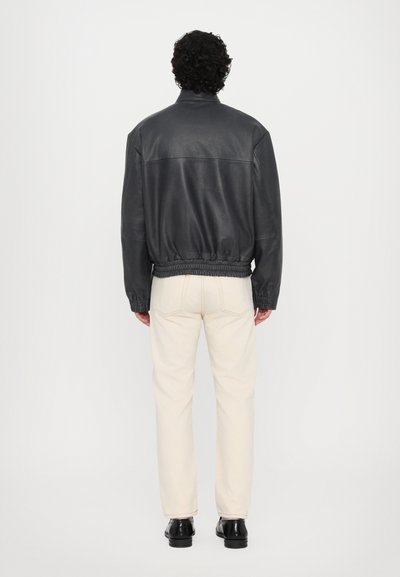 Veste bomber en cuir noir avec taille élastique et poignets, portée sur un pantalon crème et des chaussures noires, vue de dos.