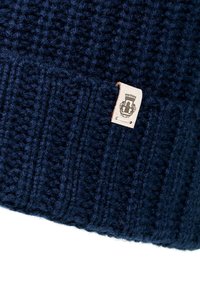 Dark blue knitted wool beanie with a small beige fabric tag featuring a black emblem stitched near the edge.