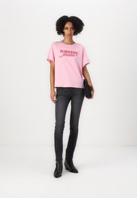 Una camiseta de manga corta rosa con el logo de "tommy jeans" en rojo. Combinada con jeans ajustados negros y botines de tacón. Diseño simple y casual.