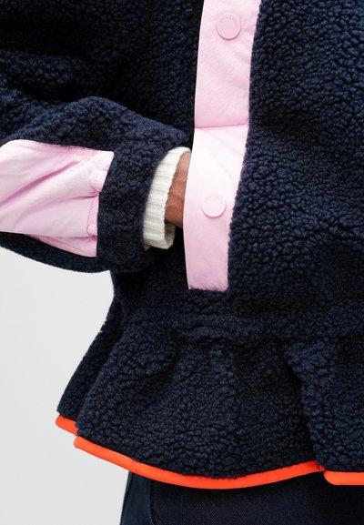 Fleece jakna z mornarsko modrim teksturo, rožnatim gumbastim ovratnikom in oranžnim detajlom ob robu; ima nabran spodnji del in slojevito zasnovo rokavov.