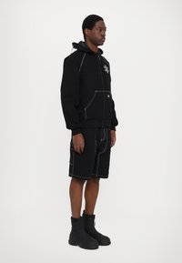 Black hoodie with contrasting white stitching, front pocket, paired with black shorts featuring similar stitching. Worn with black ankle boots.
