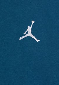 T-shirt en coton teal avec un logo Jumpman brodé en blanc. Le tissu présente une texture lisse avec des coutures minimales visibles.