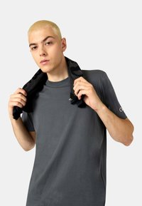 Dunkelgraues Sport-T-Shirt mit kurzen Ärmeln, glatter Textur, mit einem Logo auf dem linken Ärmel und einem schwarzen gemusterten Handtuch um den Hals drapiert.