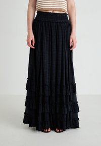 Vue de la taille vers le bas d'une personne portant une jupe maxi noire à volants avec des couches froncées et une taille smockée, associée à des sandales ouvertes.