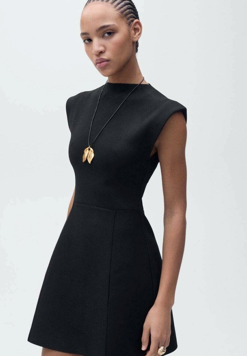 Robe noire sans manches avec un corsage ajusté et une jupe évasée, fabriquée dans un tissu lisse. Comprend un collier avec pendentif en feuille d'or.