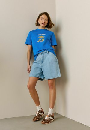 Vrouw leunend tegen een hoekmuur, gekleed in een blauw T-shirt met print, lichte denim shorts, witte sokken en sneakers met luipaardprint.