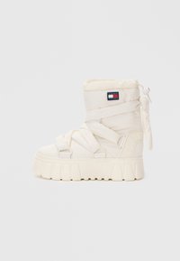 Botte blanche rembourrée avec une semelle épaisse, dotée de sangles en tissu et d'un patch logo sur le côté. Le design est à bout rond avec une texture douce.