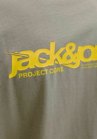 Texte jaune "jack&jones PROJECT CORE" imprimé sur un tissu gris clair, avec un texte plus petit "UNLIMITED EDITION ESTABLISHED 1990" partiellement visible.