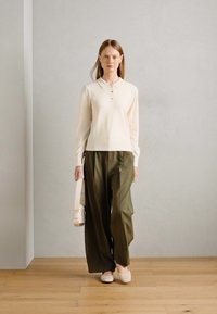 Beige langærmet polo shirt med knapper, parret med løse olivengrønne bukser og lyse beige slip-on sko, bærende en hvid taske.