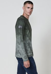 Hosszú ujjú zöld ing, texturált kamuflázs mintával, kerek nyakkivágással és KOROSHI logós patch-el a bal ujján.