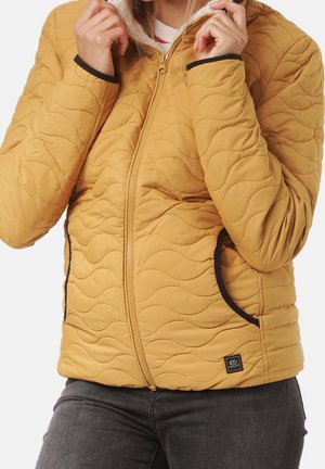 Personne portant une veste matelassée jaune avec des poches bordées de noir et une fermeture éclair à l'avant, tenant le col près du cou.