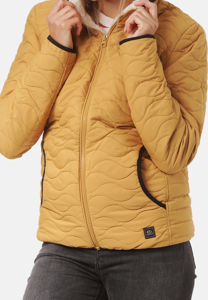 Personne portant une veste matelassée jaune avec des poches bordées de noir et une fermeture éclair à l'avant, tenant le col près du cou.