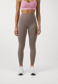 Lichtbruine hoge taille leggings met een gladde textuur, gecombineerd met een roze sport-bh met een logo-band en witte sportschoenen.