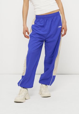 Persoană purtând pantaloni sport albastri și bej cu talie elastică și adidași albi groși, stând pe un fundal alb.