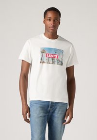 Tricou alb din bumbac cu un imprimeu grafic care înfățișează palmieri și un cer albastru, având logo-ul roșu "LEVI'S" în centru. Purtați cu blugi albaștri.