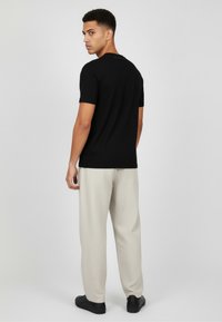 Czarny t-shirt z krótkim rękawem i okrągłym dekoltem, zestawiony z jasnobeżowymi teksturowanymi spodniami i czarnymi butami, pokazujący męski model od tyłu.