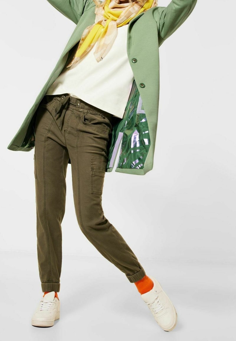 Pantaloni cargo verde oliva con polsini elastici, abbinati a un cappotto verde chiaro, una maglietta beige e una sciarpa gialla a fantasia. Sneakers bianche visibili.