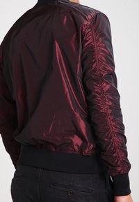 Bordowa kurtka bomberka wykonana z błyszczącego materiału, z marszczonymi rękawami i czarnym ściągaczem u dołu. Gładka faktura z minimalnymi detalami.