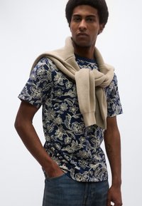 Μπλε μπλούζα με φλοράλ μοτίβο και κοντά μανίκια, συνδυασμένη με ανοιχτό μπεζ πλεκτό πουλόβερ που είναι ριγμένο στους ώμους. Ορατό τζιν παντελόνι.