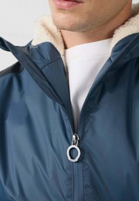 Giacca impermeabile blu con collo in pelliccia sintetica, cerniera frontale con tirante circolare, texture liscia e design minimalista.