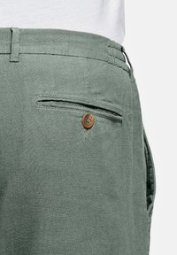 Gros plan sur une personne portant un pantalon vert clair avec une poche arrière boutonnée et une passoire de ceinture, associé à un haut blanc.