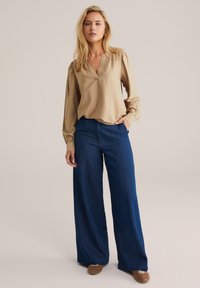 Beige Bluse mit langen Ärmeln und V-Ausschnitt, kombiniert mit weiten, dunkelblauen Jeans. Das Outfit hat eine lockere Passform und eine weiche Textur.