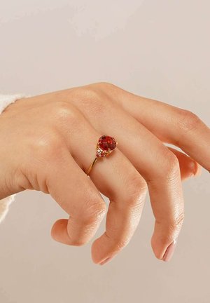Gold ring featuring a large red gemstone with a teardrop shape, accented by smaller red stones at the base, worn on a hand with light skin.