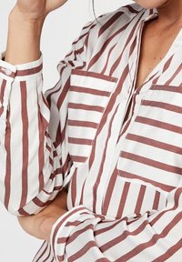 Vit och brun randig blus med krage, två framfickor och knäppte ärmslut. Bomullstyg med avslappnad passform och uppvikta ärmar.