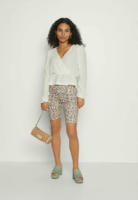 Mulher com cabelo cacheado usa uma blusa branca de mangas compridas em formato de envoltório, calções florais em tons de rosa e verde, sandálias de salto verde e segura uma bolsa cilíndrica em tom beige.