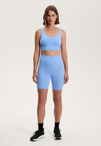 Svetlo modra športna modrc in ujemajoče visoke kolesarske kratke hlače iz raztegljivega materiala, nosene s črnimi športnimi čevlji. Minimalističen dizajn, brez vzorcev.