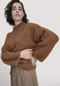 Frau mit kurzen, lockigen Haaren trägt einen groben braunen Strickpullover und lockere taupefarbene Hosen, posiert mit einer Hand auf der Hüfte.