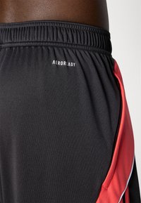 Sorte atletiske shorts lavet af åndbart stof. Har røde sideaksenter og en lynlåslomme. "AEROREADY" logo på taljen.