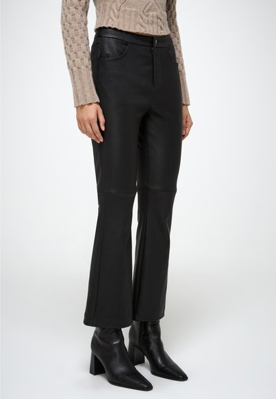 Pantalones acampanados de imitación de cuero negro con una textura suave, cierre frontal con cremallera y dos bolsillos laterales, combinados con botas de tobillo negras con punta.