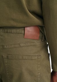 Olivgrüne Hosen mit glatter Textur, versehen mit einem braunen Lederetikett, auf dem "GANT" geprägt ist. Detail der Gesäßtasche sichtbar.
