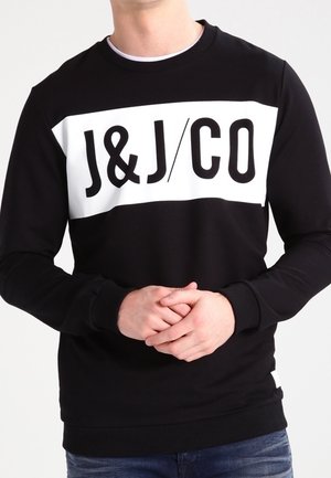 Mężczyzna w czarnej bluzie z białym prostokątnym logo z napisem "J&J/CO", z rękami złączonymi przed sobą, na jasnym, jednolitym tle.