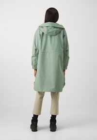 Veste imperméable légère vert menthe avec capuche, taille cintrée, poignets élastiques et poches latérales, associée à un pantalon beige coupé et des bottes noires.
