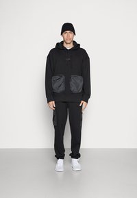 Svart hettegenser med store svarte nylonfrontlommer, sammen med svarte bukser som har sidelommer, komplettert med hvite sneakers og en lue.