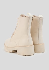 Beżowe botki za kostkę o gładkim wykończeniu, z zaokrąglonym noskiem, wiązane z przodu, z masywną podeszwą i tylnią pętlą ułatwiającą zakładanie.