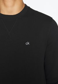 T-shirt noir à manches longues avec col rond et petit logo blanc « ck » brodé sur le côté gauche de la poitrine, porté par une personne visible du cou à la poitrine.